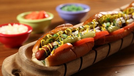 Extra long hot dog covered with toppings, and three bowls of toppings in background.
