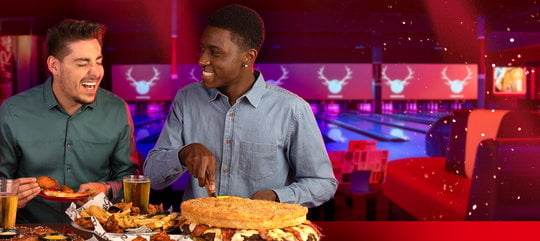 Two men eating hamburgers and chicken wings in front of bowling lanes